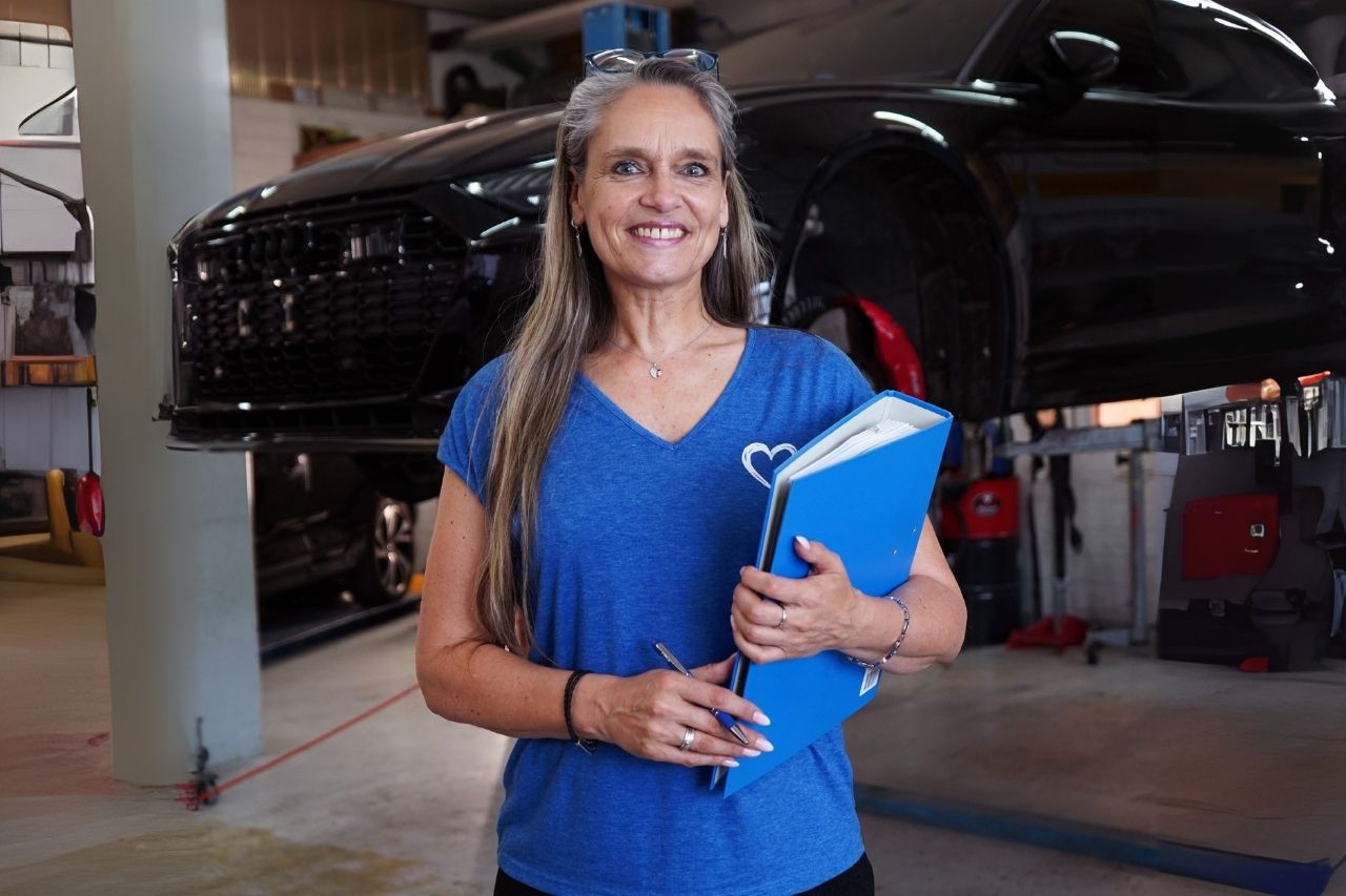 Mitarbeiterin von Lucky Cars mit blauer Mappe vor angehobenem Fahrzeug in moderner Werkstatt – kompetente Betreuung bei Autounfallreparatur, Autolackierung und Frontscheibenservice in Winterthur.